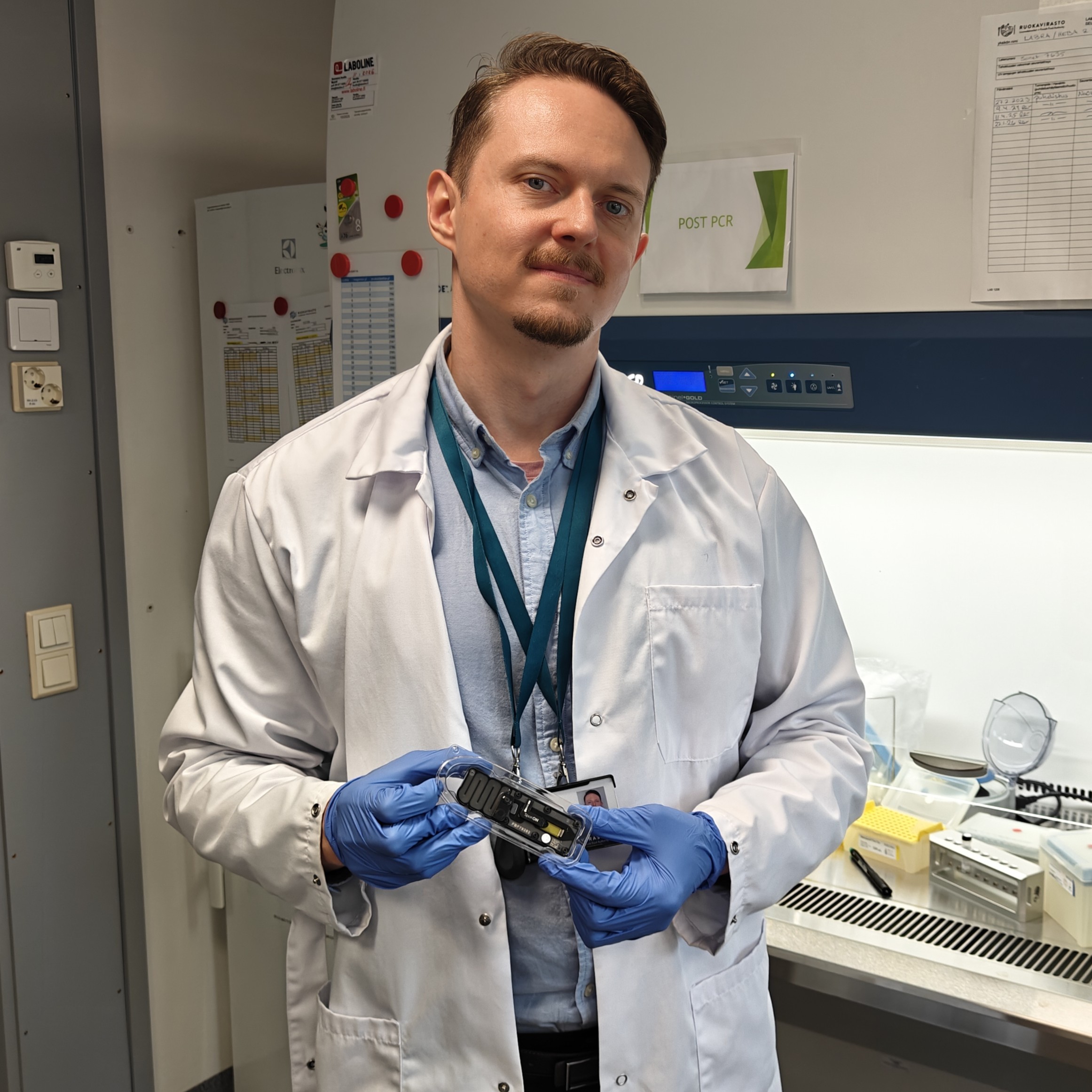 Mies valkoisessa laboratoriotakissa sinisiss&auml; kumihansikkaissa pitelee pient&auml; laitetta k&auml;siss&auml;&auml;n laboratoriossa