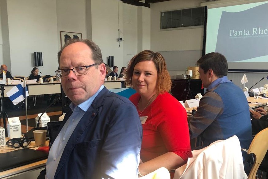 Osallistujat istuvat konferenssissa, pöydällä on Suomen lippu.
